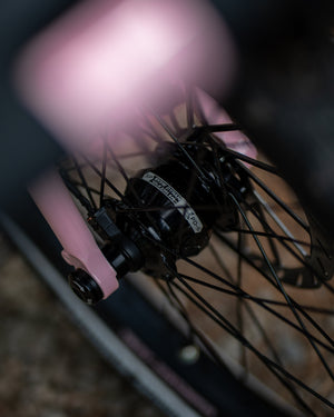 Close-up of a bicycle wheel hub and spokes with a pink frame component in the foreground and a blurred outdoor background.