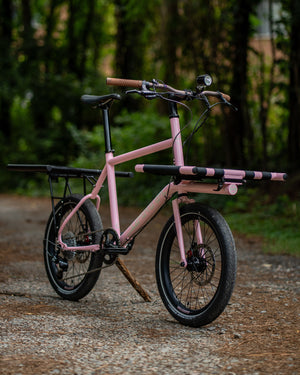 A pink utility bicycle with black components and a matching pink and black rear cargo rack, parked on a gravel path with a blurred forest background.