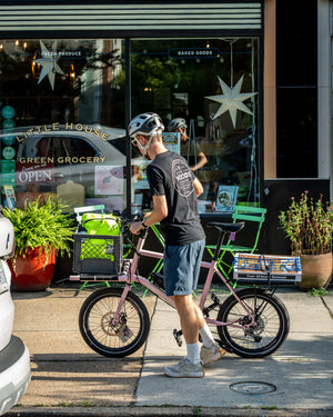 A man wearing a black t-shirt, gray shorts, and a white helmet stands next to a pink electric cargo bicycle with a front basket and rear rack on a city sidewalk in front of a storefront window.