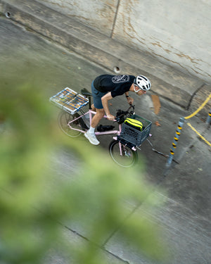 A person riding a pink cargo bicycle on a concrete path, wearing a white helmet, black t-shirt, and shorts, with a basket on the front and a box on the rear rack.
