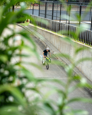 A person riding a bicycle down a concrete path outdoors, framed by blurred green foliage in the foreground and a fence along the path.