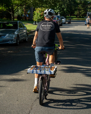 A man rides a bicycle away on a tree-lined street, wearing a black t-shirt with a white circular 'Woods & Company' logo on the back, blue shorts, and a white helmet. The bicycle has a cargo rack carrying a box strapped down with blue tape.