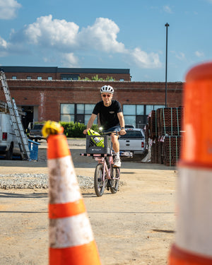 A person riding a bicycle in an outdoor construction area, wearing a white helmet and sunglasses, with orange traffic cones in the foreground and a brick building in the background.