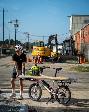 A man wearing a white helmet and black t-shirt stands next to a pink cargo bicycle with a wooden board strapped to its rear rack, set in an outdoor construction site with heavy machinery in the background.